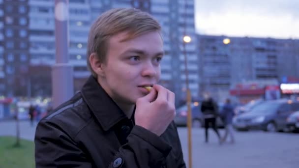 Kille äter pommes frites potatis på gatan, på bänken. Mannen äter pommes frites, stekt potatis, chip, ohälsosamma skräp snabbmat. ung man äta en burgare med stekt potatis i gata mat café. Snabbmat — Stockvideo