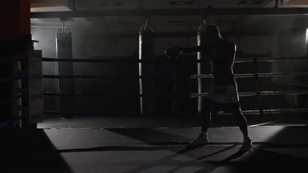 Кикбокс истребитель тень бокса на ринге. Спортсмен борется со своей тенью. Молодой боксер на тренировке наносит удар бинтами на кулаки, пока тренируется на ринге — стоковое фото