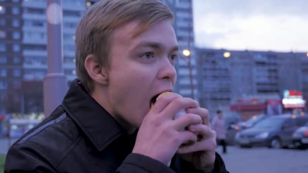 若い男が通りのハンバーガーでおいしいファーストフードのハンバーガーを噛んで食べること。若い男が路上でハンバーガーを食べる — ストック動画