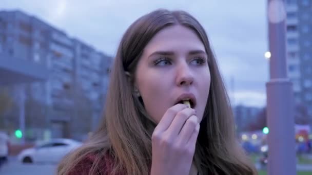 En ung flicka har middag snabbmat på torget. Ung kvinna stående i en urban street och äter pommes frites. Ung kvinna äta snabbmat stående på gatan — Stockvideo