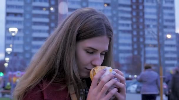 Mujer joven parada en una calle urbana y comiendo hamburguesa. Mujer joven comiendo comida rápida de pie en la calle — Vídeo de stock