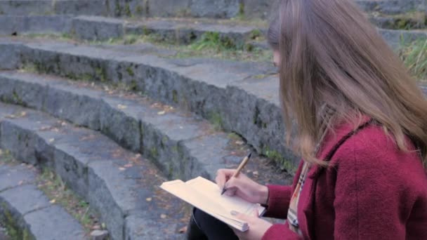 Atractiva joven estudiante que se prepara para dar conferencias en la Universidad mientras está sentada en las escaleras, escribiendo notas en su cuaderno de notas. Chica joven con un cuaderno en la mano — Vídeo de stock