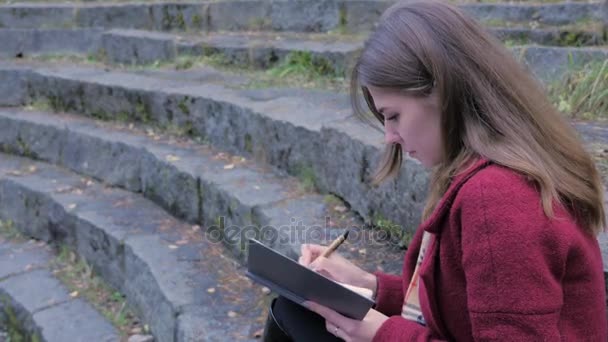 Jovem estudante atraente se preparando para palestras na Universidade enquanto se senta nas escadas, escrevendo notas em seu livro. Jovem com um caderno na mão — Vídeo de Stock