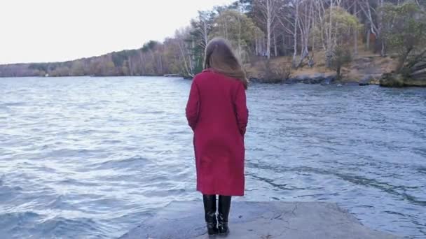 Chica joven con el pelo se para cerca del agua en la estación fría. Vista trasera. Mujer joven de pie cerca de un lago — Vídeos de Stock