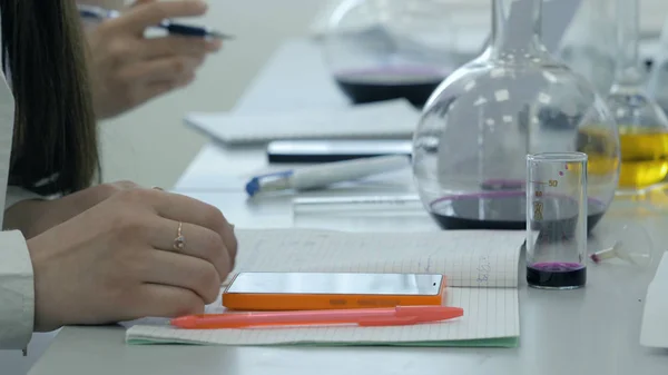 Os estudantes no trabalho no laboratório da química tomam notas em um caderno. Aluno feminino usando telefone na aula de ciências. Jovens estudantes de medicina escrever — Fotografia de Stock