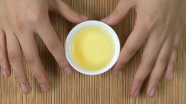 Mãos femininas segurando xícara branca de chá chinês fundo de bambu, vista superior. Uma mulher a segurar uma chávena de chá. Chá sencha japonês verde em flores caneca — Fotografia de Stock