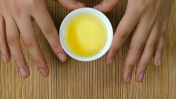 Womans kezei fehér csésze tea kínai bambusz háttér, felülnézet. A nő, kezében egy csésze teát. Zöld japán sencha teát virágok bögre — Stock Fotó