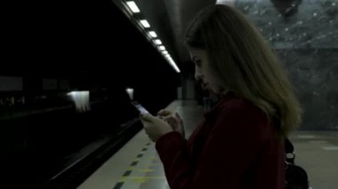 Kız Metro İstasyonu'na ve tren için bekler, akıllı telefon veya tablet kullanarak kırmızı paltolu. Kadın kullanın cep telefonu ve ayakta şehir metro staton.