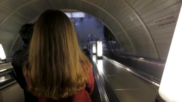 Vista trasera de una chica bajando por la escalera mecánica en el metro. Mujer bajando a la estación de metro a través de la escalera mecánica tounel. mujer montando una escalera mecánica en una estación de metro — Vídeos de Stock