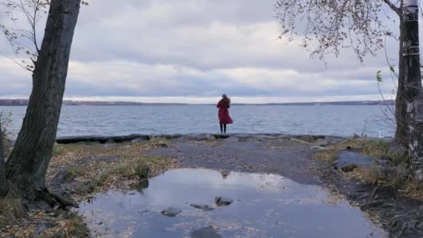 Z tyłu widok dziewczyny z płynącej czerwone włosy ubrany w czerwony płaszcz na tle jezioro i autumn drzewa. Piękna dziewczyna kaukaski spędzania czasu nad jeziorem, w lesie. Kobieta relaksujący w lesie. — Wideo stockowe