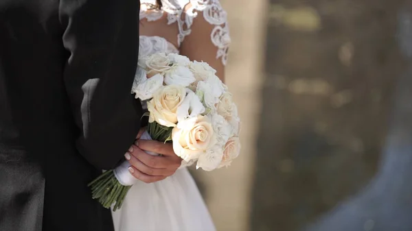 Elegante Braut und Bräutigam posieren zusammen im Freien an einem Hochzeitstag. Braut hält einen weißen Rosenstrauß, während sie neben dem Bräutigam steht. Braut in einem Kleid stehend und mit einem Brautstrauß — Stockfoto