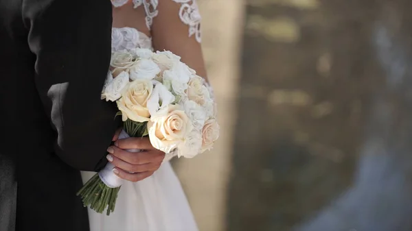 Elegante Braut und Bräutigam posieren zusammen im Freien an einem Hochzeitstag. Braut hält einen weißen Rosenstrauß, während sie neben dem Bräutigam steht. Braut in einem Kleid stehend und mit einem Brautstrauß — Stockfoto