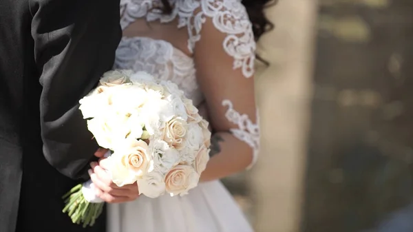 Elegante Braut und Bräutigam posieren zusammen im Freien an einem Hochzeitstag. Braut hält einen weißen Rosenstrauß, während sie neben dem Bräutigam steht. Braut in einem Kleid stehend und mit einem Brautstrauß — Stockfoto