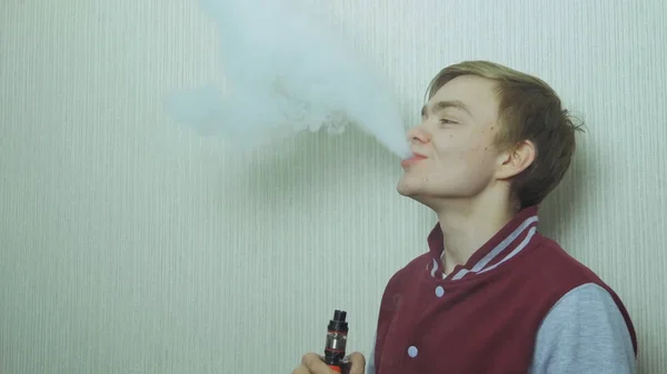 Fiatal férfi vaping egy elektronikus cigaretta. Vaper füst csípő párologtató. A fiatalembert élvezi egy kielégítő e-cigaretta állandó profil, fehér falhoz — Stock Fotó