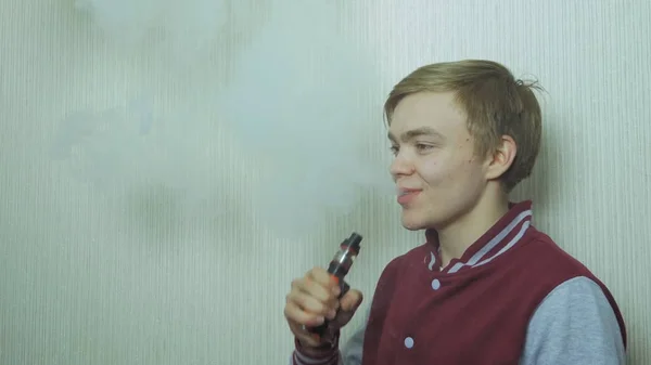 Un giovanotto che fuma una sigaretta elettronica. Vaper vaporizzatore di fumo hipster. Giovane uomo godendo di una soddisfacente sigaretta elettronica in piedi di profilo contro parete bianca — Foto Stock