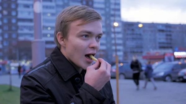 Kille äter pommes frites potatis på gatan, på bänken. Mannen äter pommes frites, stekt potatis, chip, ohälsosamma skräp snabbmat. ung man äta en burgare med stekt potatis i gata mat café. Snabbmat — Stockfoto