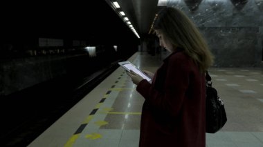 Kız Metro İstasyonu'na ve tren için bekler, akıllı telefon veya tablet kullanarak kırmızı paltolu. Kadın kullanın cep telefonu ve ayakta şehir metro staton.