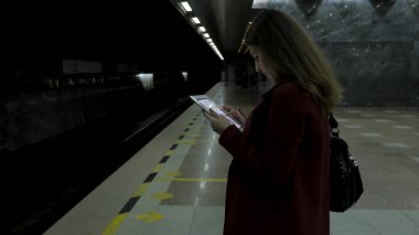 Kız Metro İstasyonu'na ve tren için bekler, akıllı telefon veya tablet kullanarak kırmızı paltolu. Kadın kullanın cep telefonu ve ayakta şehir metro staton.