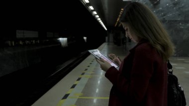 Kız Metro İstasyonu'na ve tren için bekler, akıllı telefon veya tablet kullanarak kırmızı paltolu. Kadın kullanın cep telefonu ve ayakta şehir metro staton.