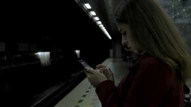 Kız Metro İstasyonu'na ve tren için bekler, akıllı telefon veya tablet kullanarak kırmızı paltolu. Kadın kullanın cep telefonu ve ayakta şehir metro staton.