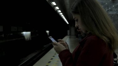 Kız Metro İstasyonu'na ve tren için bekler, akıllı telefon veya tablet kullanarak kırmızı paltolu. Kadın kullanın cep telefonu ve ayakta şehir metro staton.