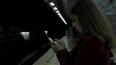 Kız Metro İstasyonu'na ve tren için bekler, akıllı telefon veya tablet kullanarak kırmızı paltolu. Kadın kullanın cep telefonu ve ayakta şehir metro staton.