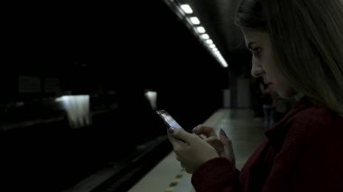 Kız Metro İstasyonu'na ve tren için bekler, akıllı telefon veya tablet kullanarak kırmızı paltolu. Kadın kullanın cep telefonu ve ayakta şehir metro staton.