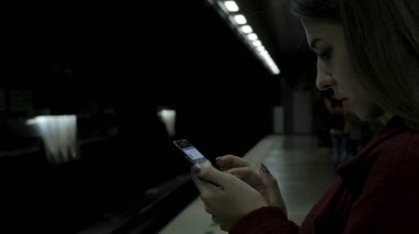 Kız Metro İstasyonu'na ve tren için bekler, akıllı telefon veya tablet kullanarak kırmızı paltolu. Kadın kullanın cep telefonu ve ayakta şehir metro staton.
