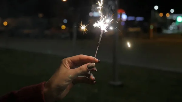 Beautiful sparkler in woman hand on black or street background. Woman holding sparkler against colorful defocused lights, close up. Female hands holding sparkler on dark background — Stock Photo, Image