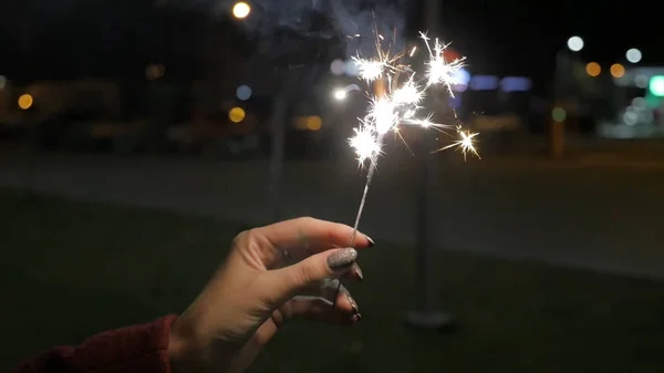 Beautiful sparkler in woman hand on black or street background. Woman holding sparkler against colorful defocused lights, close up. Female hands holding sparkler on dark background — Stock Photo, Image