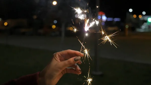 Lindo sparkler na mão mulher no fundo preto ou rua. Mulher segurando sparkler contra luzes coloridas desfocadas, de perto. Mãos femininas segurando sparkler no fundo escuro — Fotografia de Stock