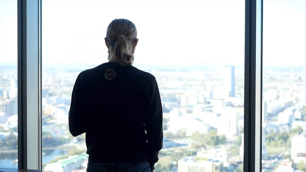 Rückansicht junger erfolgreicher Geschäftsmann, der im Büro steht und durch ein Fenster auf die Stadtlandschaft mit Wolkenkratzern blickt, träumt oder rastet, auf Besprechungen wartet oder Geschäftsangebote in Betracht zieht. — Stockfoto