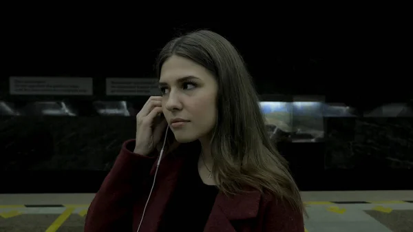 若い女の子は、地下鉄トンネル内のヘッドフォンで音楽を聴きます。地下鉄トンネル内の大きなヘッドホンで音楽を聴く若い女子学生。若い女性で彼女のスマート フォンで音楽を聴き、 — ストック写真