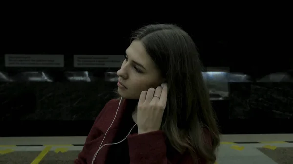 Молодая девушка слушает музыку в наушниках в туннеле метро. Молодая студентка слушает музыку в больших наушниках в тоннеле метро. Молодая женщина слушает музыку на своем смартфоне в — стоковое фото