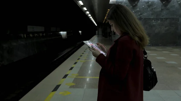 Chica de abrigo rojo usando smartphone o tableta en la estación de metro y espera al tren. Mujer uso de teléfono celular y de pie en la ciudad de metro staton . — Foto de Stock