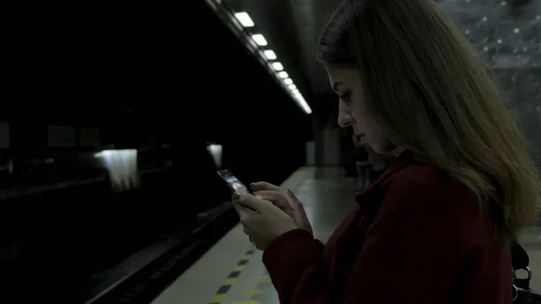 Девушка в красном пальто, используя смартфон или планшет на станции метро и ждет поезда. Пользование сотовым телефоном и пребывание в городском метро . — стоковое фото