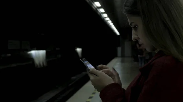 Девушка в красном пальто, используя смартфон или планшет на станции метро и ждет поезда. Пользование сотовым телефоном и пребывание в городском метро . — стоковое фото