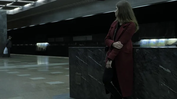 Ładna dziewczyna z Czapka słuchanie muzyki w stacji metra. Kobieta za pomocą jej telefon komórkowy w metrze, ona słucha muzyki za pomocą słuchawek. Ciemny Portret młodej kobiety w metrze — Zdjęcie stockowe