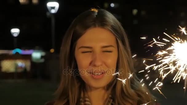 Menina adolescente sorridente na rua à noite com faíscas. Jovem celebrando um evento o Ano Novo está chegando. Retrato de menina segurando faíscas em câmera lenta — Vídeo de Stock