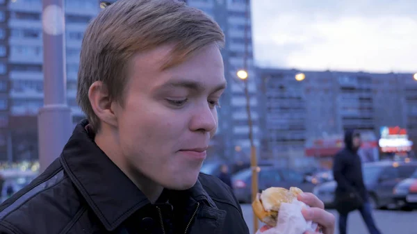 若い男が通りのハンバーガーでおいしいファーストフードのハンバーガーを噛んで食べること。若い男が路上でハンバーガーを食べる — ストック写真