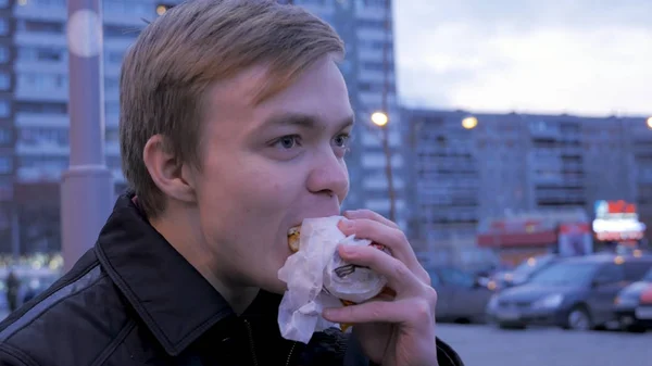 吃在街头汉堡咬一个可口的快餐食品汉堡包的年轻男子。年轻人在街上吃一个汉堡 — 图库照片