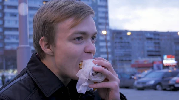 Nuori mies syö maukasta pikaruokahampurilaista katupurilaisella. Nuori mies syö hampurilaista kadulla. — kuvapankkivalokuva