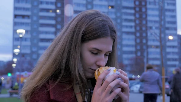Nuori nainen seisoo urbaanilla kadulla ja syö hampurilaisia. Nuori nainen syö pikaruokaa seisoo kadulla — kuvapankkivalokuva