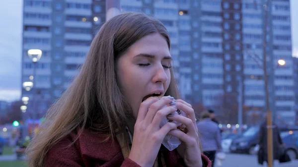 Young woman standing in an urban street and eating burger. Young woman eating fast food standing on the street — Stock Photo, Image