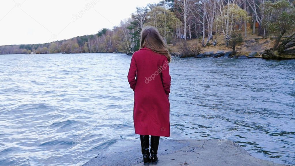 Young girl with her hair stands near the water in the cold season. Back view. Young woman standing near a lake