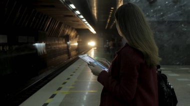 Kız Metro İstasyonu'na ve tren için bekler, akıllı telefon veya tablet kullanarak kırmızı paltolu. Kadın kullanın cep telefonu ve ayakta şehir metro staton.