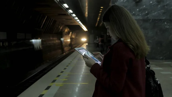 Дівчина в червоному пальто використовує смартфон або планшет на станції метро і чекає на поїзд. Жінка використовує мобільний телефон і стоїть в стадоні міського метро . — стокове фото