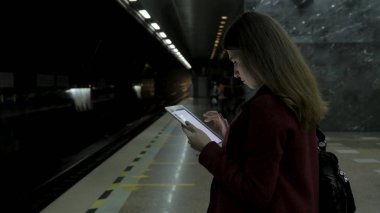 Kız Metro İstasyonu'na ve tren için bekler, akıllı telefon veya tablet kullanarak kırmızı paltolu. Kadın kullanın cep telefonu ve ayakta şehir metro staton.