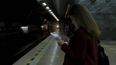 Kız Metro İstasyonu'na ve tren için bekler, akıllı telefon veya tablet kullanarak kırmızı paltolu. Kadın kullanın cep telefonu ve ayakta şehir metro staton.