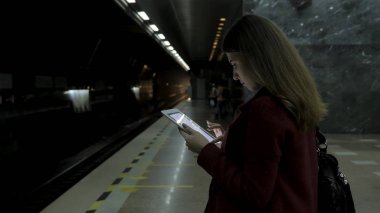 Kız Metro İstasyonu'na ve tren için bekler, akıllı telefon veya tablet kullanarak kırmızı paltolu. Kadın kullanın cep telefonu ve ayakta şehir metro staton.